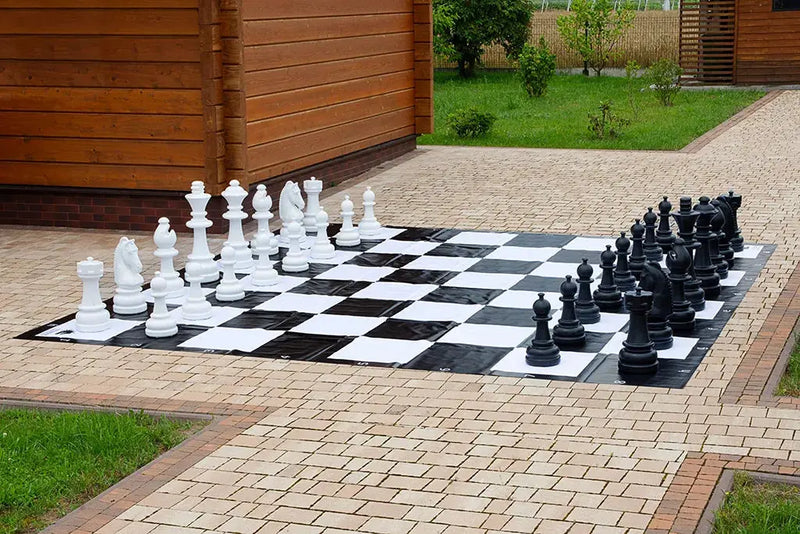 Pièces d'échecs géantes en plastique, blanches et noires, sur un échiquier géant posé sur une terrasse pavée devant un chalet en bois